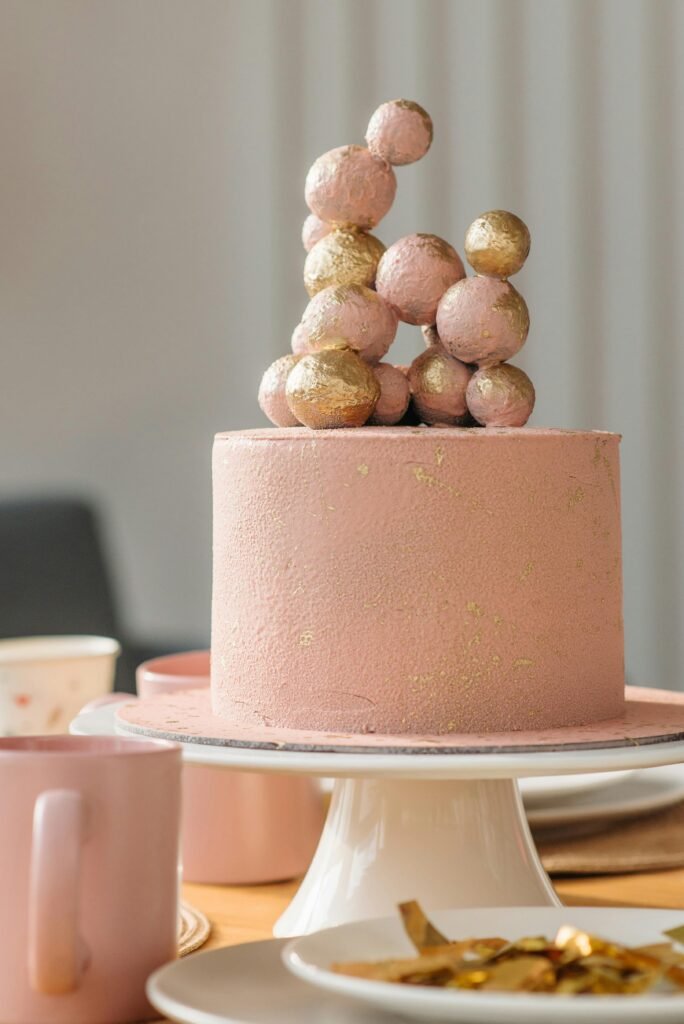 Pink cake topped with gold spheres, elegant design on a table with plates and mugs.